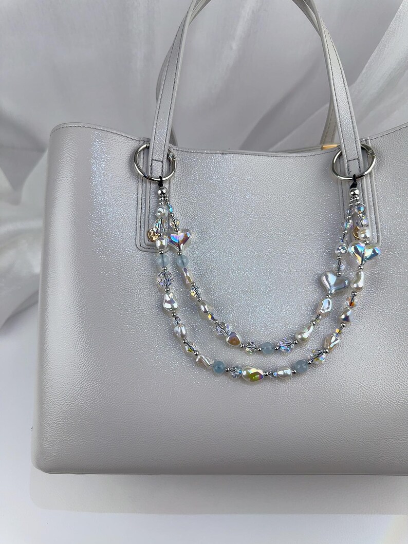 May include: A light gray tote bag with silver hardware and handles. The bag is adorned with two beaded charms featuring iridescent beads, pearls, and heart-shaped accents. The bag has a glossy finish.