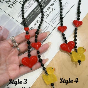 May include: Two necklaces with black beads and red heart charms. The necklace on the left has a large red heart charm and a yellow rubber duck charm. The necklace on the right has a smaller red heart charm and a yellow rubber duck charm. Both necklaces are 17 inches long. The text "Style 3 Custom big heart 17 in" is below the necklace on the left. The text "Style 4 Custom big heart 17 in" is below the necklace on the right.