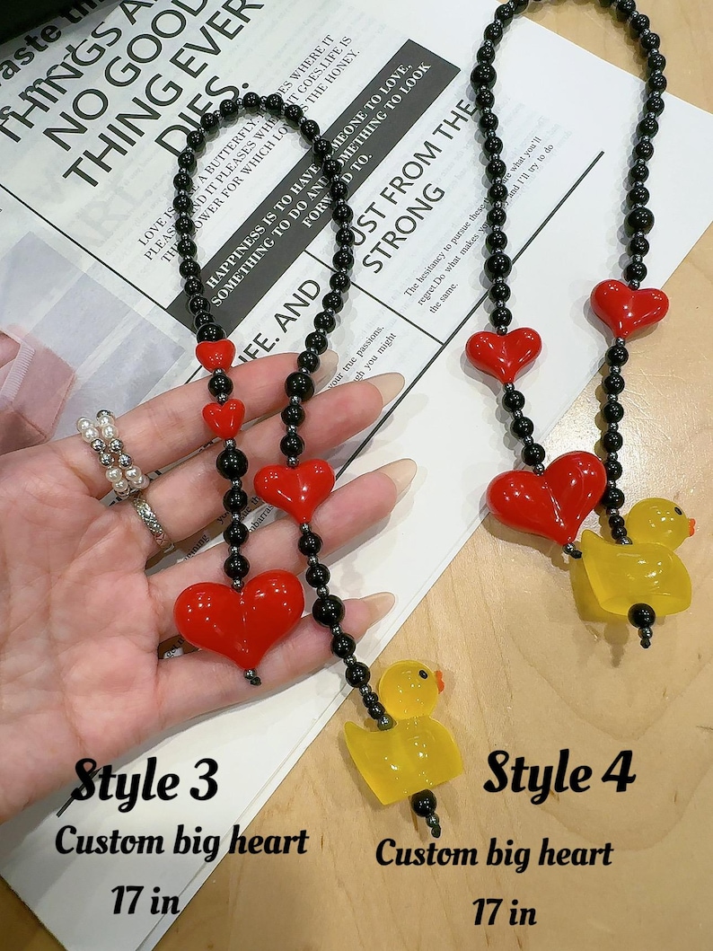 May include: Two necklaces with black beads and red heart charms. The necklace on the left has a large red heart charm and a yellow rubber duck charm. The necklace on the right has a smaller red heart charm and a yellow rubber duck charm. Both necklaces are 17 inches long. The text "Style 3 Custom big heart 17 in" is below the necklace on the left. The text "Style 4 Custom big heart 17 in" is below the necklace on the right.