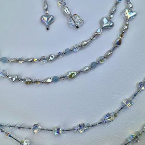 May include: A multi-strand beaded accessory featuring clear faceted crystals, iridescent heart-shaped beads, and light blue round beads. The strands are connected to a silver-toned metal ring, with dangling segments of beads ending in heart and crystal charms.