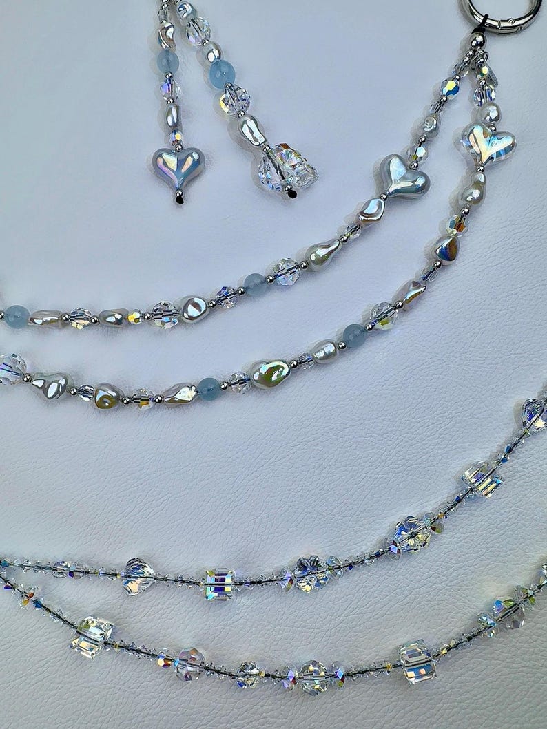 May include: A multi-strand beaded accessory featuring clear faceted crystals, iridescent heart-shaped beads, and light blue round beads. The strands are connected to a silver-toned metal ring, with dangling segments of beads ending in heart and crystal charms.