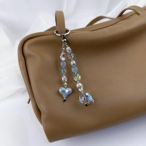 May include: A bag charm featuring two dangling strands of beads attached to a silver-toned metal ring. The strands include iridescent heart-shaped beads, clear faceted crystals, and white pearls in varying sizes, hanging from a tan leather bag.