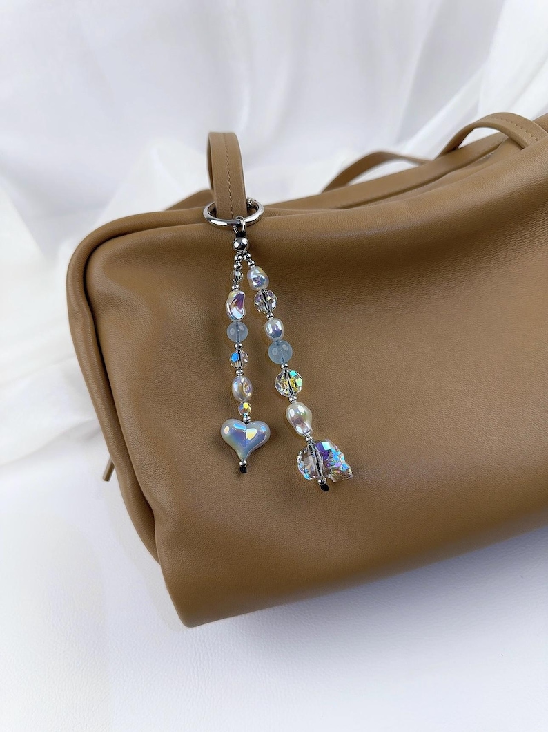 May include: A bag charm featuring two dangling strands of beads attached to a silver-toned metal ring. The strands include iridescent heart-shaped beads, clear faceted crystals, and white pearls in varying sizes, hanging from a tan leather bag.