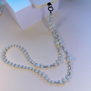 May include: A white phone case with a pearl and crystal phone strap. The strap is attached to the case with a black metal ring.