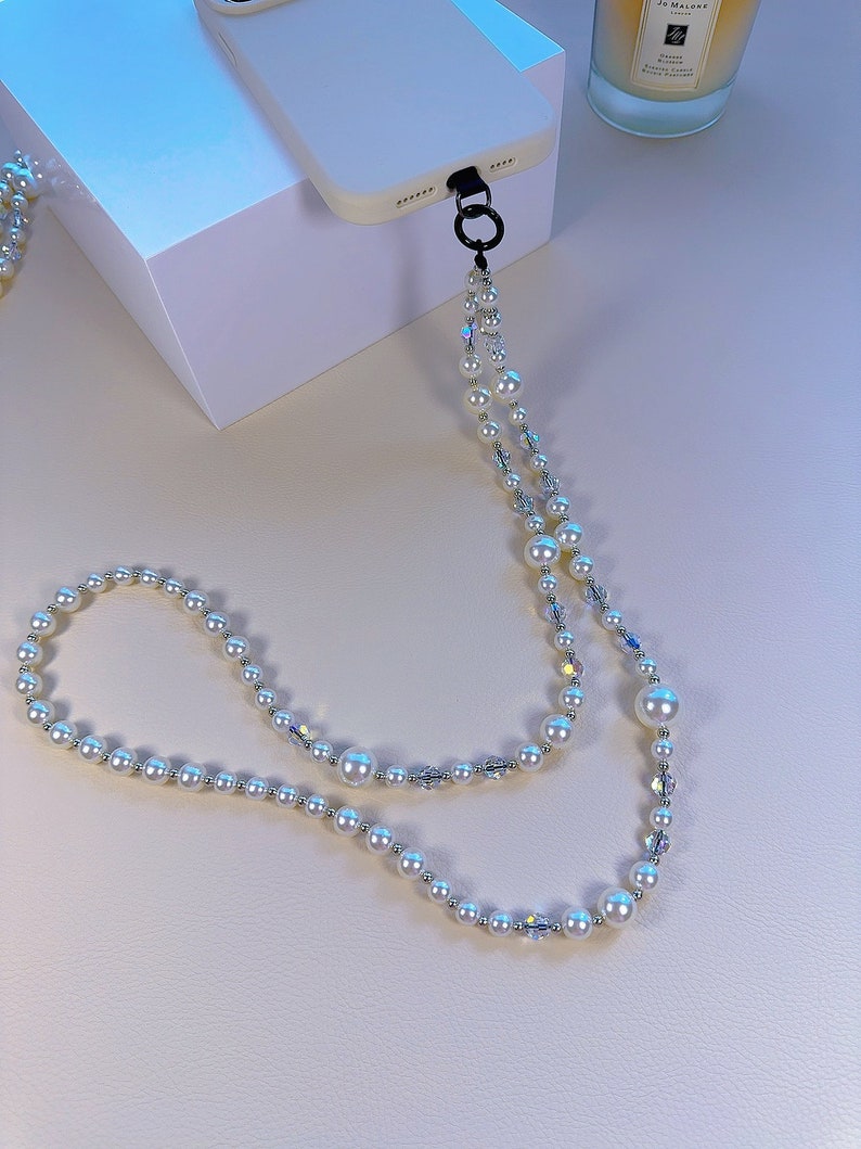 May include: A white phone case with a pearl and crystal phone strap. The strap is attached to the case with a black metal ring.