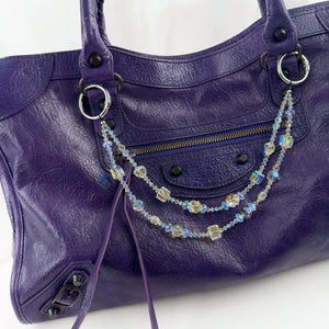 May include: A deep purple leather handbag featuring a textured surface, dual handles, and a front zippered pocket. A decorative double-strand chain made of iridescent, multi-faceted crystal beads is attached to the silver-toned metal rings.