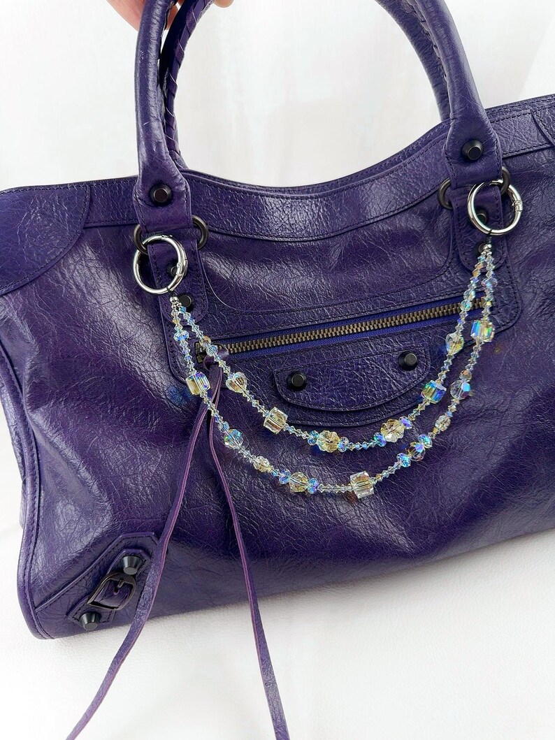 May include: A deep purple leather handbag featuring a textured surface, dual handles, and a front zippered pocket. A decorative double-strand chain made of iridescent, multi-faceted crystal beads is attached to the silver-toned metal rings.