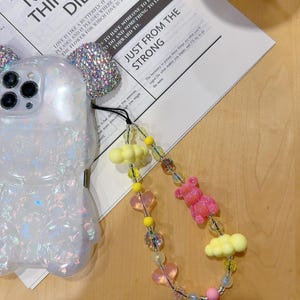 May include: A colourful phone charm featuring pink and yellow heart-shaped beads, yellow cloud charms, and a pink bear charm. The charm is attached to a white phone case with a sparkly design.