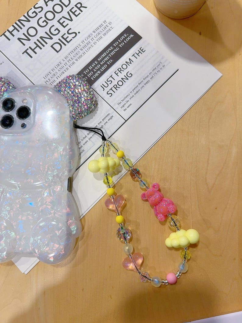 May include: A colourful phone charm featuring pink and yellow heart-shaped beads, yellow cloud charms, and a pink bear charm. The charm is attached to a white phone case with a sparkly design.