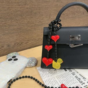 May include: A black leather handbag with a black and red beaded chain with red heart charms and a yellow rubber duck charm. The bag has a silver clasp and a top handle.