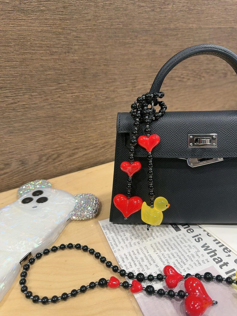 May include: A black leather handbag with a black and red beaded chain with red heart charms and a yellow rubber duck charm. The bag has a silver clasp and a top handle.