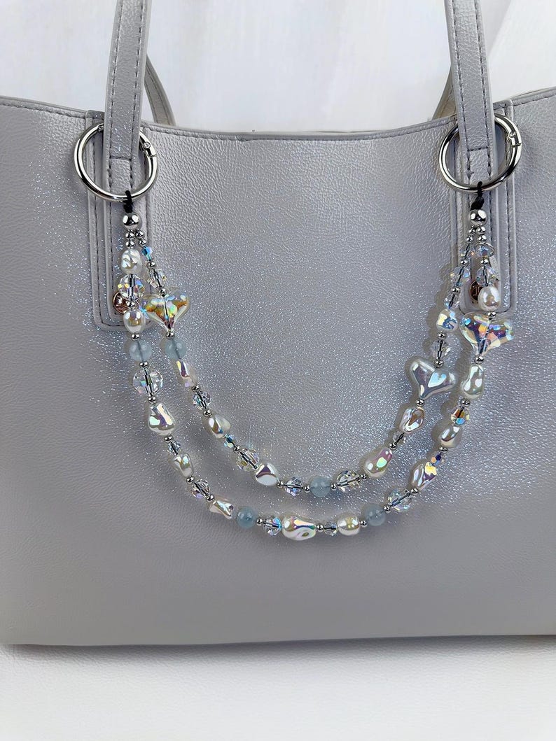 May include: A decorative double-strand bag charm featuring iridescent heart-shaped beads, white baroque pearls, clear faceted crystals, and small light blue round beads. The charm is attached to a light gray handbag with silver-toned metal rings.