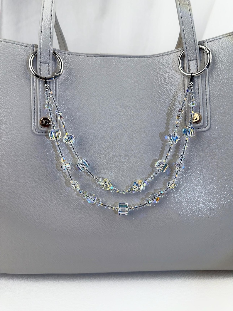 May include: A light gray handbag with silver hardware and a beaded strap. The strap is made of clear, iridescent beads in various shapes, strung on a black cord. The bag has two silver rings for attaching the strap.