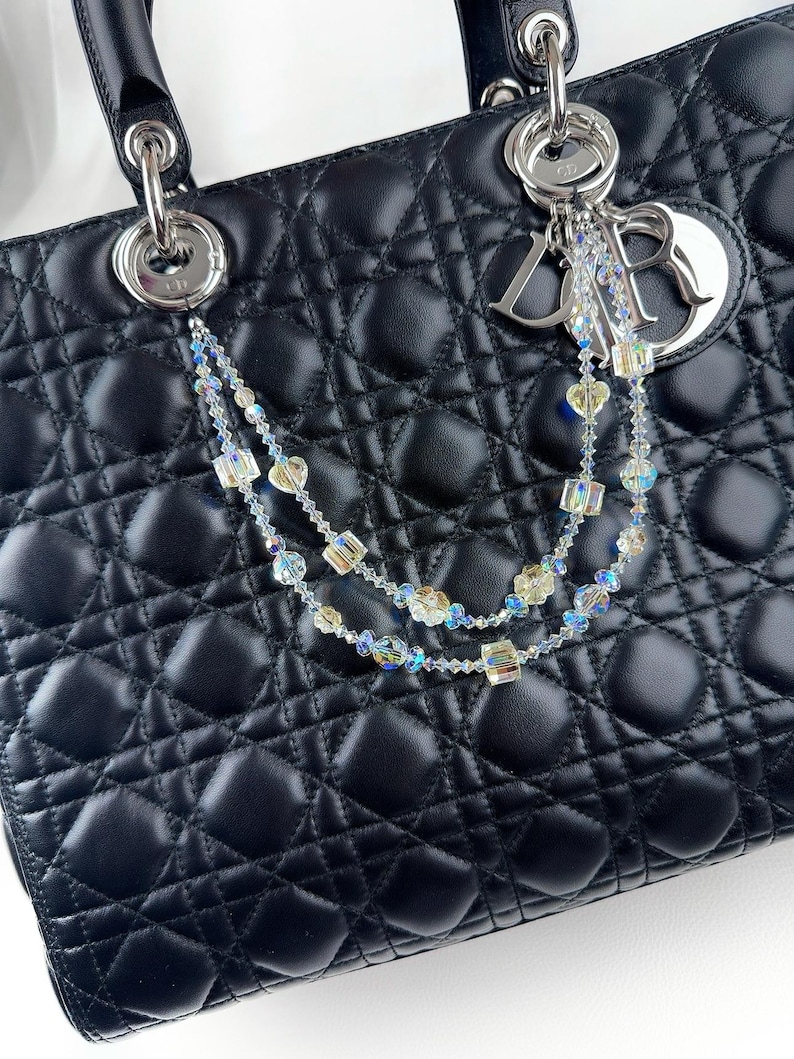 May include: A black quilted leather handbag with silver hardware and a decorative crystal beaded charm. The charm features iridescent beads and a silver logo tag. The bag has a top handle and a diamond pattern.
