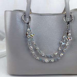 May include: A light gray tote bag featuring a decorative double-strand beaded chain attached to the silver-toned rings. The chain consists of iridescent heart-shaped beads, clear faceted crystals, and small light blue round beads.