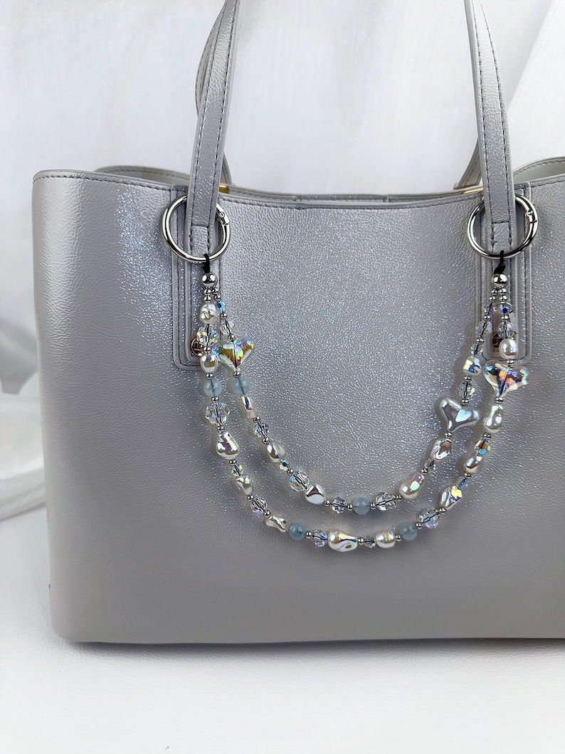 May include: A light gray tote bag featuring a decorative double-strand beaded chain attached to the silver-toned rings. The chain consists of iridescent heart-shaped beads, clear faceted crystals, and small light blue round beads.