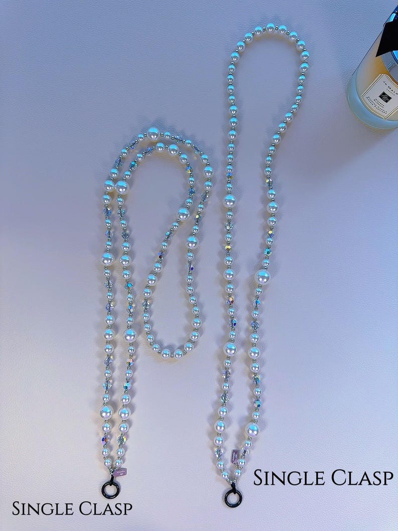 May include: Two white pearl and crystal beaded phone straps with single clasps. The straps are on a white background.  SINGLE CLASP is written on the bottom right corner of the image.