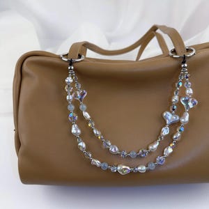 May include: A tan leather handbag featuring a decorative double-strand beaded chain attached to silver-toned rings. The chain consists of iridescent heart-shaped beads, white baroque pearls, small clear faceted crystals, and smooth light blue beads.