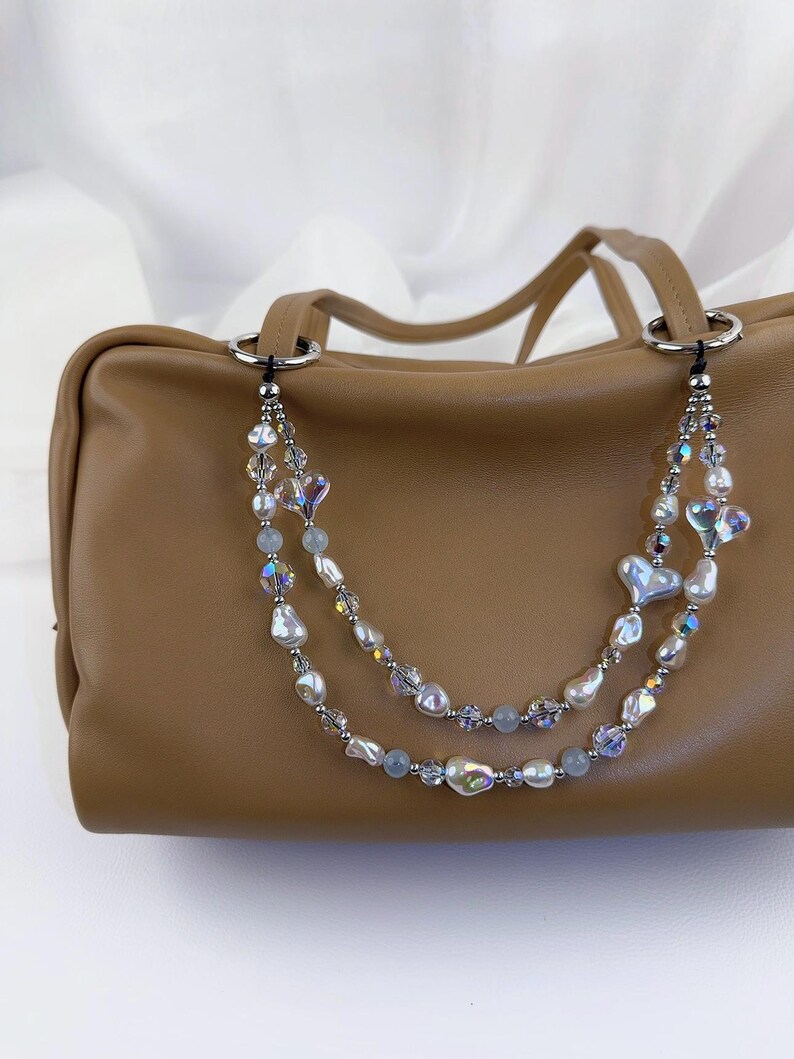 May include: A tan leather handbag featuring a decorative double-strand beaded chain attached to silver-toned rings. The chain consists of iridescent heart-shaped beads, white baroque pearls, small clear faceted crystals, and smooth light blue beads.