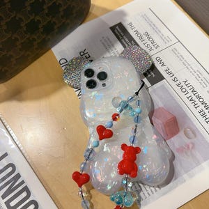 May include: A clear phone case with a sparkly iridescent finish and a 3D bear design. The case has a beaded chain with red hearts and blue beads attached to it. The case is designed for an iPhone.