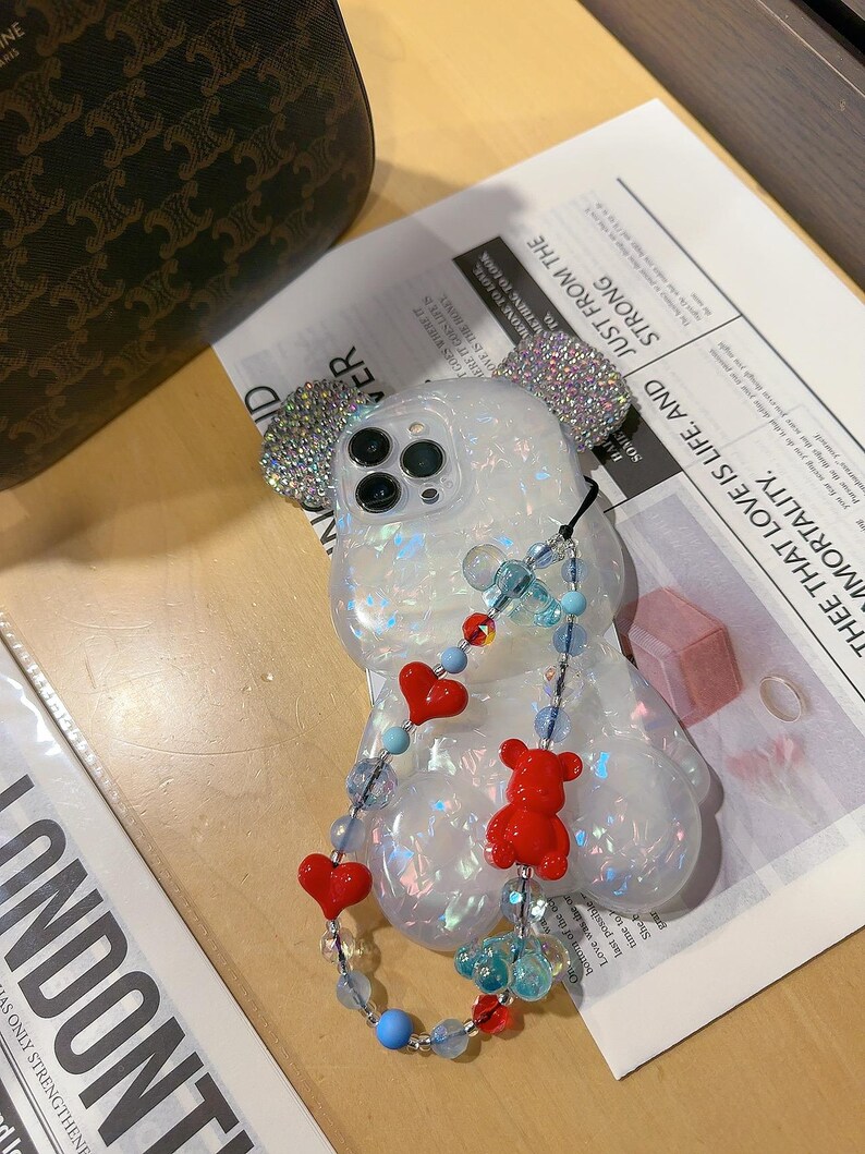 May include: A clear phone case with a sparkly iridescent finish and a 3D bear design. The case has a beaded chain with red hearts and blue beads attached to it. The case is designed for an iPhone.