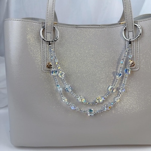 May include: A light gray tote bag featuring a decorative double-strand beaded chain accessory. The chain is composed of clear, iridescent, and cube-shaped crystals attached to the bag's silver-toned metal rings with small black clasps.