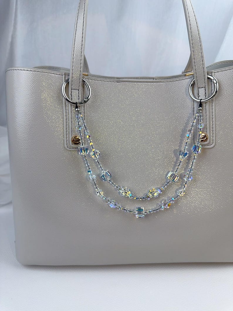 May include: A light gray tote bag featuring a decorative double-strand beaded chain accessory. The chain is composed of clear, iridescent, and cube-shaped crystals attached to the bag's silver-toned metal rings with small black clasps.