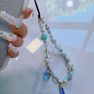 May include: A white and iridescent beaded phone charm with a crystal pendant. The charm features a mix of white pearls, iridescent beads, and a blue and green iridescent bead.