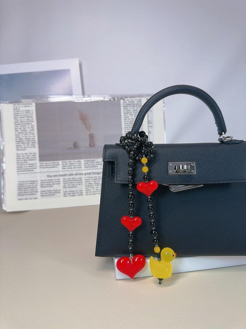 May include: A black leather handbag with a gold chain and a black beaded charm with red heart and yellow duck accents.