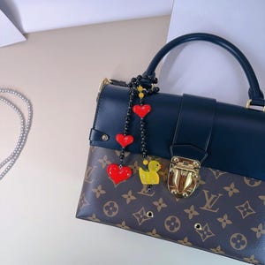 May include: A black leather handbag with a brown and tan monogram pattern. The bag has a gold clasp and a black beaded strap with red heart and yellow duck charms.
