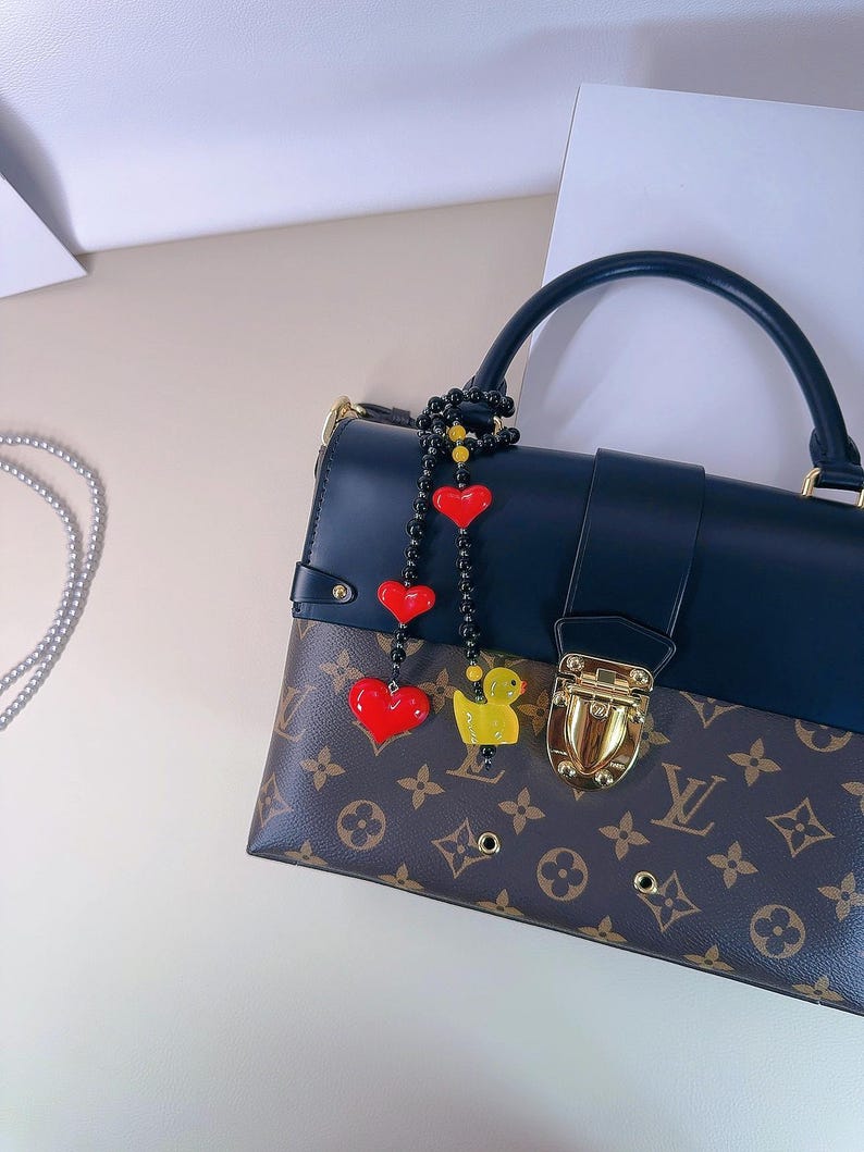 May include: A black leather handbag with a brown and tan monogram pattern. The bag has a gold clasp and a black beaded strap with red heart and yellow duck charms.