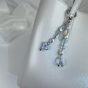 May include: A silver-toned metal bag charm featuring two dangling strands of beads attached to a circular ring. The beads include iridescent heart-shaped charms, clear faceted crystals, light blue spheres, and small white pearls.