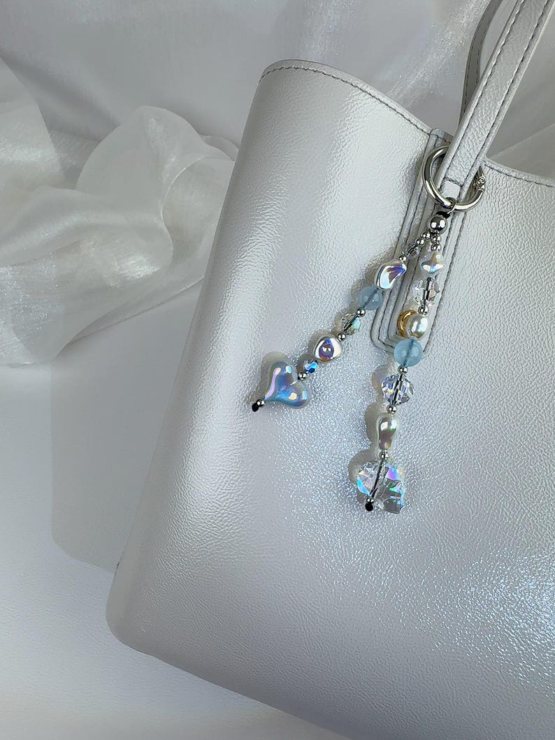 May include: A silver-toned metal bag charm featuring two dangling strands of beads attached to a circular ring. The beads include iridescent heart-shaped charms, clear faceted crystals, light blue spheres, and small white pearls.