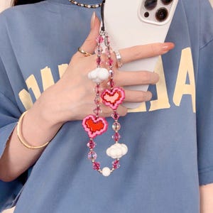 May include: A phone charm with pink and red heart and cloud charms. The charm is strung with pink and clear beads. The phone case is white. The person is wearing a blue t-shirt and gold jewellery.