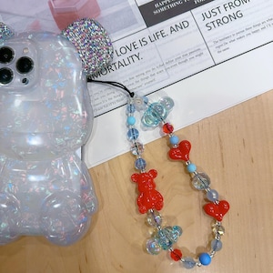 May include: A clear phone case with a sparkly iridescent finish shaped like a bear with a beaded phone charm. The charm features red heart beads, red bear beads, and blue and clear glass beads. The charm is attached to the phone case with a silver ring. The phone case is sitting on a white surface with text that reads "LOVE IS LIKE A BUTTERFLY... THE FLOWER FOR WHICH LOVE IS THE HOME", "HAPPINESS IS TO HAVE SOMEONE TO LOVE... SOMETHING TO DO AND SOMETHING TO LOOK FORWARD TO", "LOVE IS LIFE. AND JUST FROM THE STRONG", and "The hesitancy to pursue these things are what regret. Do what makes you happy and I'll do the same."
