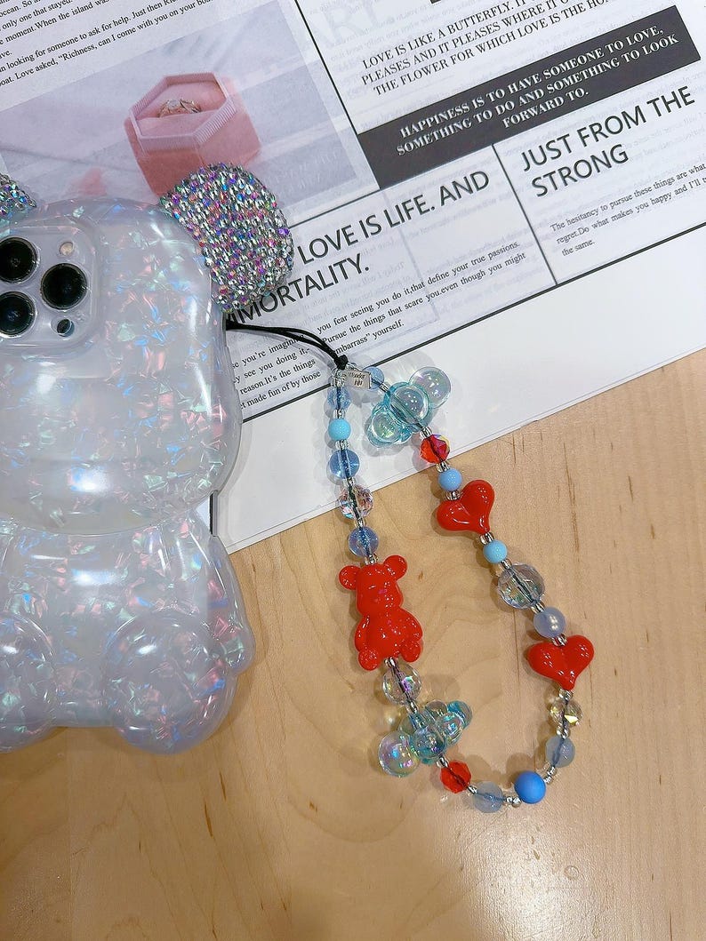 May include: A clear phone case with a sparkly iridescent finish shaped like a bear with a beaded phone charm. The charm features red heart beads, red bear beads, and blue and clear glass beads. The charm is attached to the phone case with a silver ring. The phone case is sitting on a white surface with text that reads "LOVE IS LIKE A BUTTERFLY... THE FLOWER FOR WHICH LOVE IS THE HOME", "HAPPINESS IS TO HAVE SOMEONE TO LOVE... SOMETHING TO DO AND SOMETHING TO LOOK FORWARD TO", "LOVE IS LIFE. AND JUST FROM THE STRONG", and "The hesitancy to pursue these things are what regret. Do what makes you happy and I'll do the same."