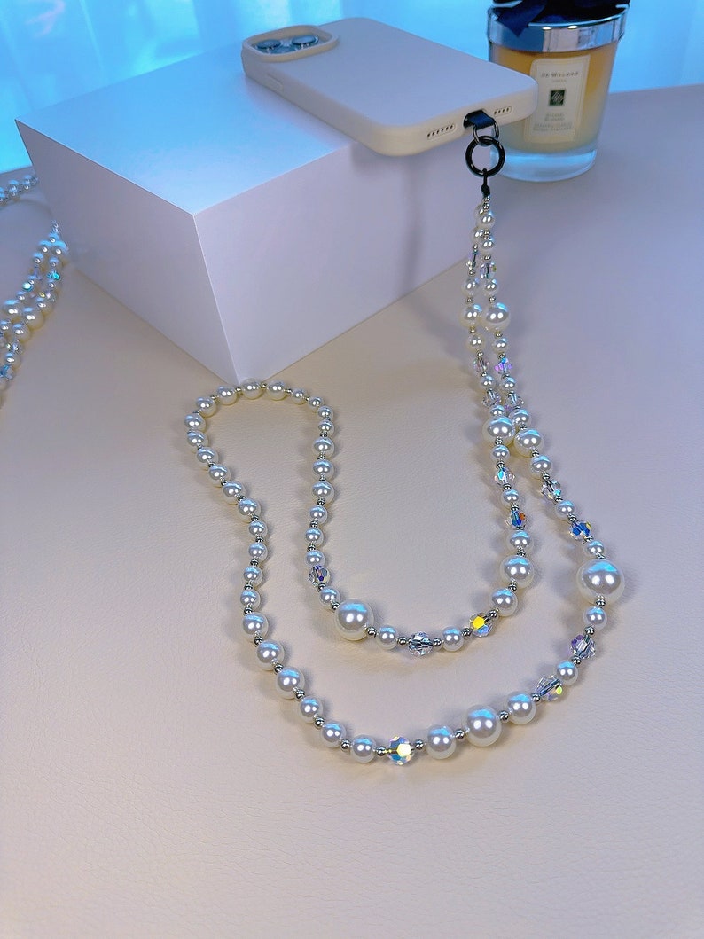 May include: A white phone case with a pearl and crystal phone strap. The strap is made of two strands of pearls and crystals.