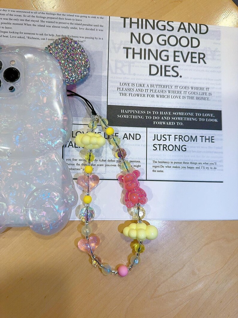May include: A pink and yellow beaded phone charm with a pink bear, yellow clouds, and pink hearts. The charm is hanging from a phone case. The text on the card reads "Things and no good thing ever dies. Love is like a butterfly. It goes where it pleases and it pleases where it goes. Life is the flower for which love is the honey. Happiness is to have someone to love, something to do and something to look forward to. Just from the strong."