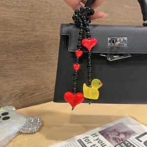 May include: A black leather handbag with a black beaded chain strap with red heart charms and a yellow rubber duck charm. The text "If you love for time." is visible in the background.