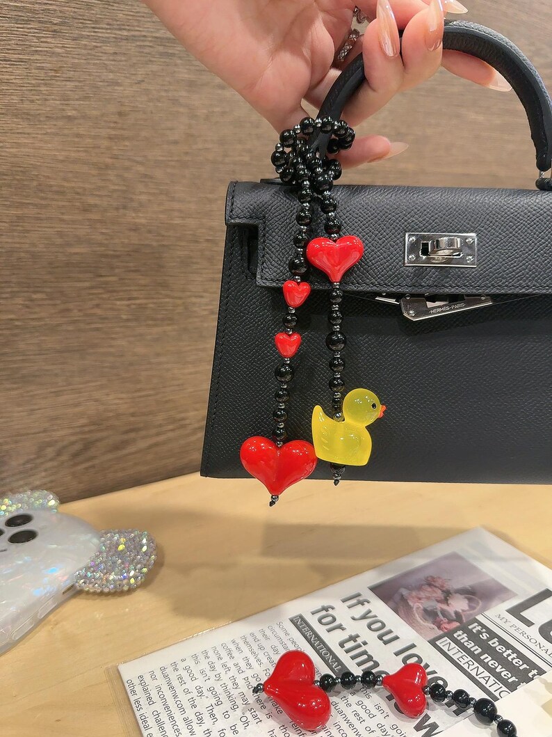 May include: A black leather handbag with a black beaded chain strap with red heart charms and a yellow rubber duck charm. The text "If you love for time." is visible in the background.