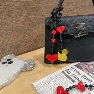 May include: A black leather handbag with a silver clasp and a red heart charm attached to a black beaded chain. The chain also has a yellow rubber duck charm. The handbag is sitting on a wooden surface with a white phone case with a cartoon ghost design.