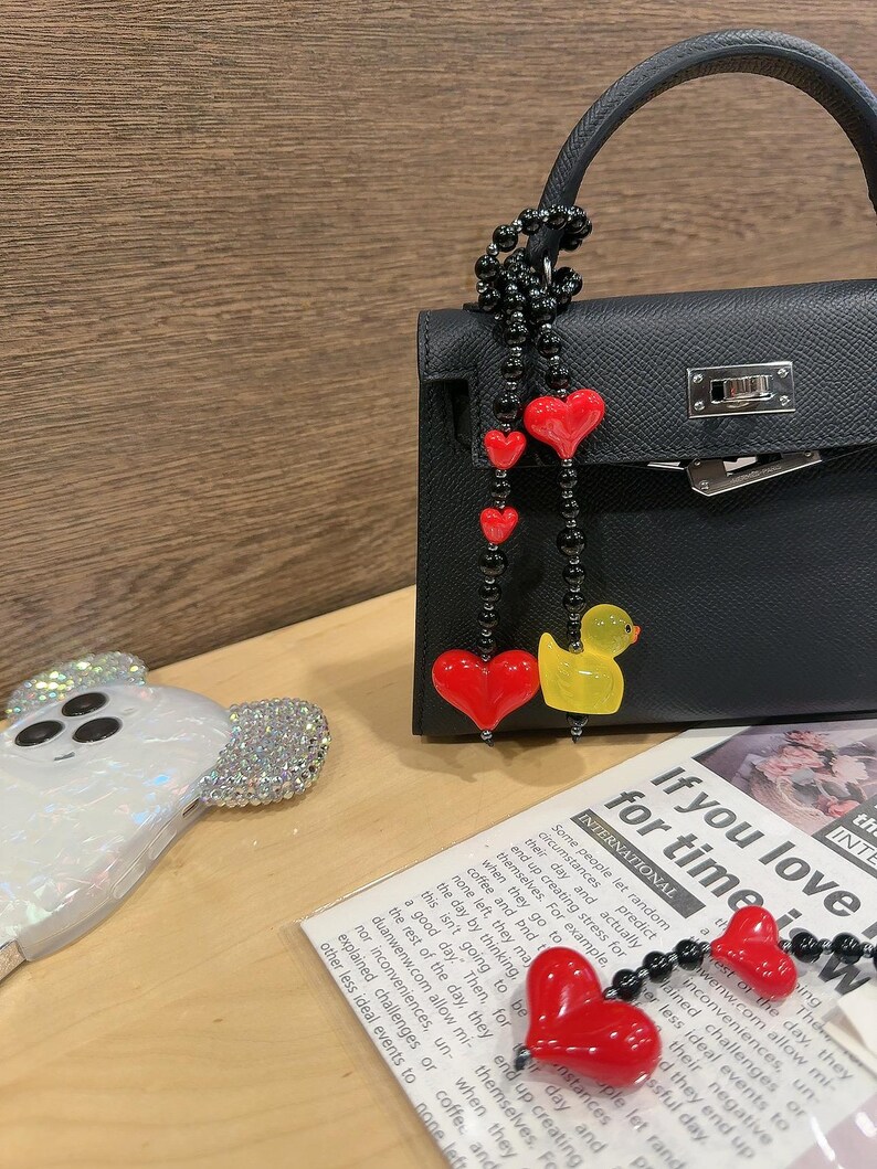 May include: A black leather handbag with a silver clasp and a red heart charm attached to a black beaded chain. The chain also has a yellow rubber duck charm. The handbag is sitting on a wooden surface with a white phone case with a cartoon ghost design.