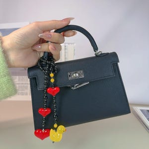 May include: A black leather handbag with a top handle and a gold clasp. A black beaded chain with red heart charms and a yellow rubber duck charm hangs from the handle.