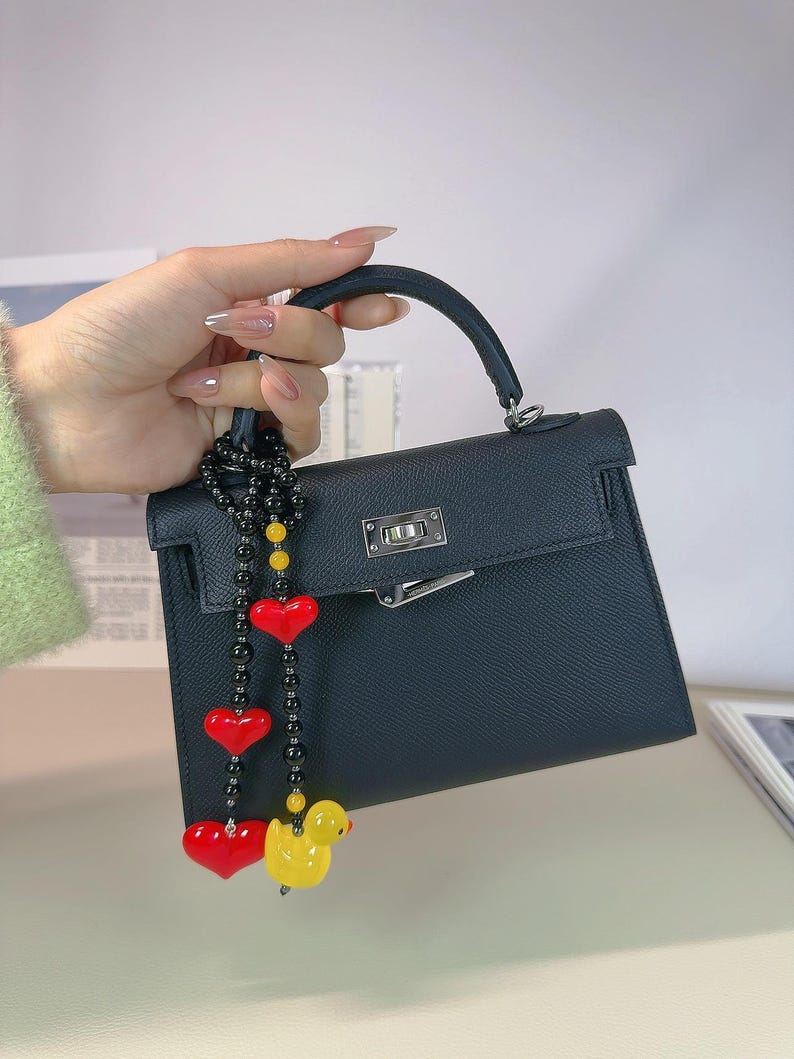 May include: A black leather handbag with a top handle and a gold clasp. A black beaded chain with red heart charms and a yellow rubber duck charm hangs from the handle.