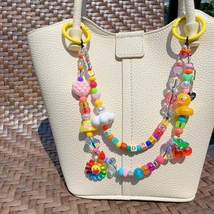 Könnte beinhalten: Eine cremefarbene, strukturierte Handtasche mit einem dekorativen Perlenriemen, der an zwei gelben Ringen befestigt ist. Der Riemen enthält bunte Perlen, eine gelbe Gummiente, eine weiße Wolke, einen Pilz, Kirschen und eine Blume mit dem Text SUNDAY.