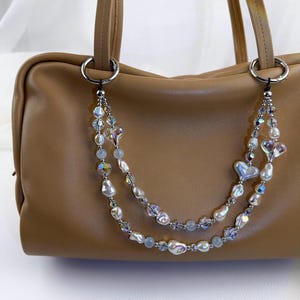 May include: A tan leather handbag featuring a decorative double-strand beaded chain attached to silver-toned rings. The chain consists of iridescent crystal beads, white baroque pearls, and heart-shaped charms in a layered, dangling arrangement.