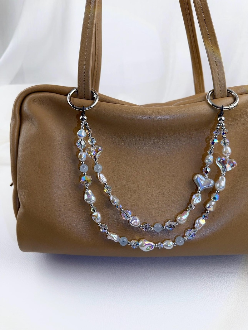 May include: A tan leather handbag featuring a decorative double-strand beaded chain attached to silver-toned rings. The chain consists of iridescent crystal beads, white baroque pearls, and heart-shaped charms in a layered, dangling arrangement.
