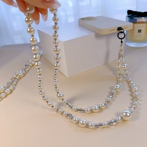 May include: A white pearl and crystal phone strap with a silver clasp. The strap is made of two strands of pearls and crystals.