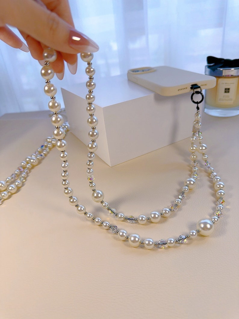 May include: A white pearl and crystal phone strap with a silver clasp. The strap is made of two strands of pearls and crystals.