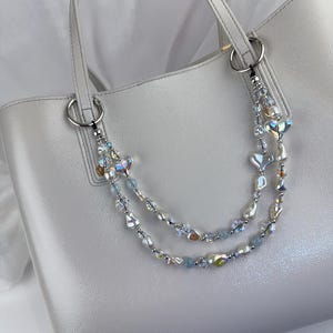 May include: A light gray tote bag featuring a decorative double-strand beaded chain attached to the silver-toned rings. The chain consists of iridescent heart-shaped beads, white faux pearls, small clear crystals, and light blue round beads.